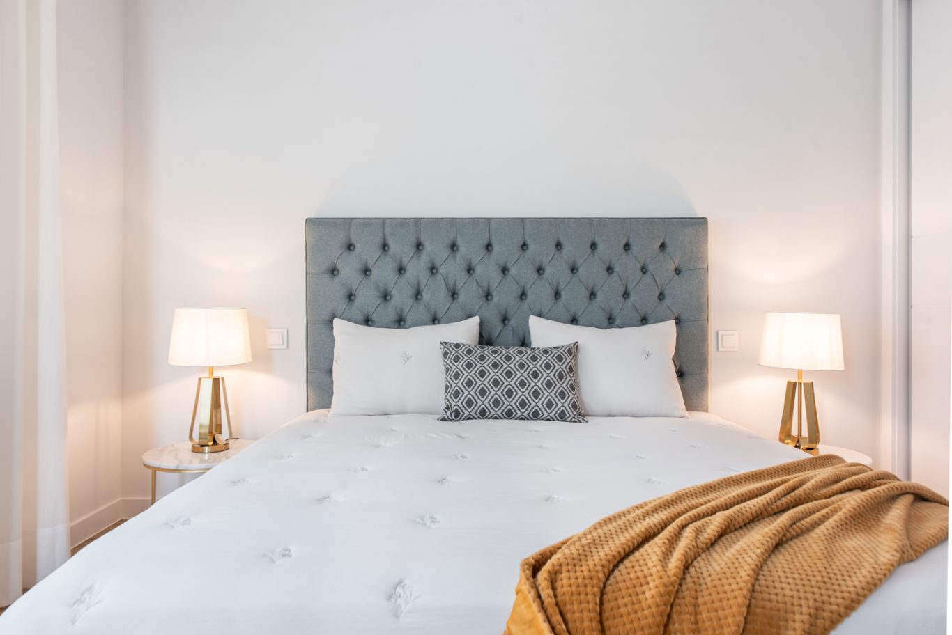 Cama con cabecera gris, almohadas blancas y manta amarilla, iluminada por lámparas.